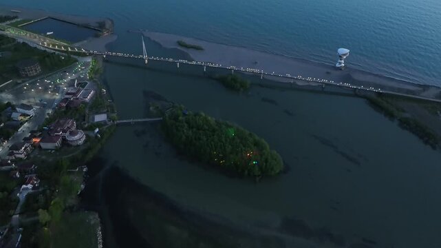 Horizontal drone aerial footage frame capturing the long wooden pedestrian bridge in Anaklia, showcasing the river delta meeting the Black Sea beach, a small green island, and coastal resort