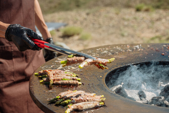 Close-up of a chef in black gloves cooking bacon-wrapped asparagus on a large round outdoor grill with a central fire pit