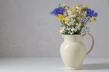 Fototapeta premium Beautiful wildflowers in jug on white wooden table near grey wall, closeup. Space for text