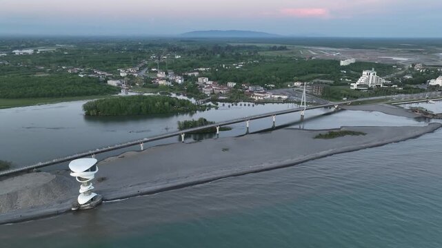 Horizontal drone aerial footage frame capturing the long wooden pedestrian bridge in Anaklia, showcasing the river delta meeting the Black Sea beach, a small green island, and coastal resort