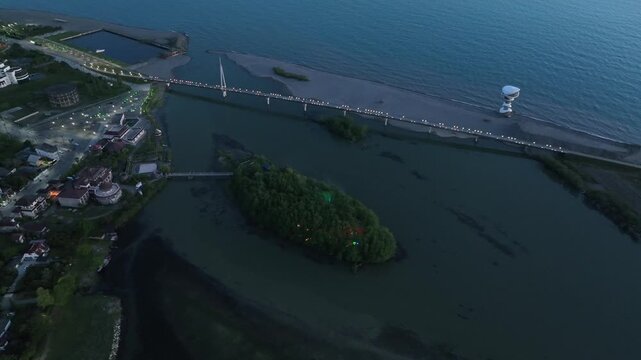 Horizontal drone aerial footage frame capturing the long wooden pedestrian bridge in Anaklia, showcasing the river delta meeting the Black Sea beach, a small green island, and coastal resort