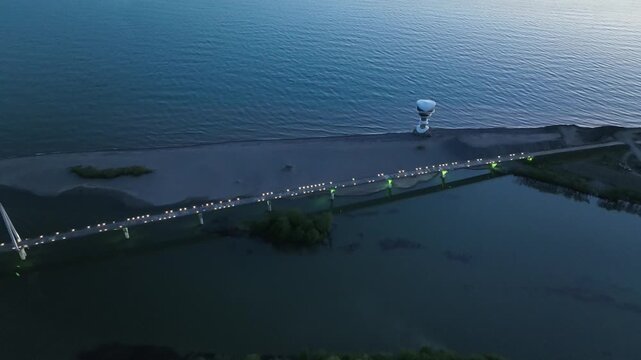 Horizontal drone aerial footage frame capturing the long wooden pedestrian bridge in Anaklia, showcasing the river delta meeting the Black Sea beach, a small green island, and coastal resort