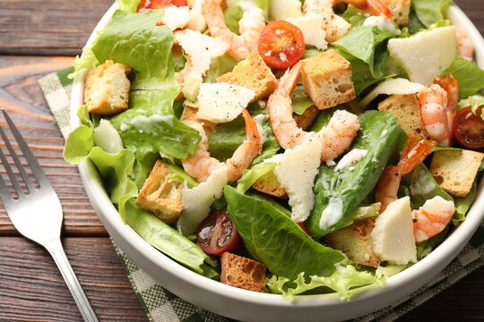 Delicious Caesar salad with shrimps in bowl served on wooden table, closeup