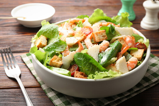 Delicious Caesar salad with shrimps in bowl served on wooden table, closeup