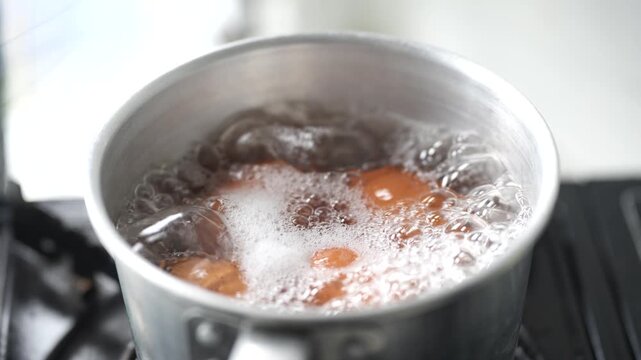 Four brown chicken eggs are boiling in a stainless steel pot filled with hot water. The close-up image captures bubbling water and steam during cooking, highlighting a simple kitchen preparation