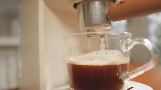 A close-up coffee maker brewing hot coffee into a clear glass mug, releasing a fragrant aroma and a smooth, inviting foam in the warm morning light.