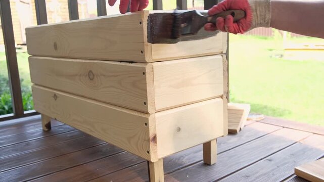 Brown oil for protection of wood by staining. Painting fence for protecting and pests. Woodworker applying finish to pine board box. Repairman varnishing with brush wooden fencing wall plank surface.