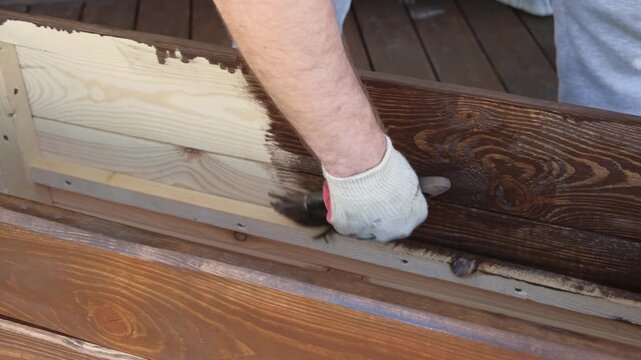 Brown oil for protection of wood by staining. Painting fence for protecting and pests. Woodworker applying finish to pine board box. Repairman varnishing with brush wooden fencing wall plank surface.