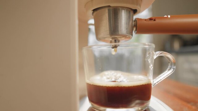 A close-up coffee maker brewing hot coffee into a clear glass mug, releasing a fragrant aroma and a smooth, inviting foam in the warm morning light.