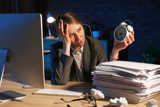 Workaholic. Overwhelmed woman with alarm clock, stack of documents and computer at wooden desk indoors at night