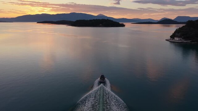 Beautiful drone footage following a lone boat across calm waters during a golden sunrise in Lefkada, Greece. The vessel leaves an elegant wake pattern behind as warm morning light r