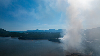 Obraz premium Aerial view of a landfill fire in Phuket with heavy smoke spreading across nearby landscapes and urban areas. Thick plumes of smoke rise illustrating the impact of urban waste and air pollution.