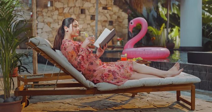 Indian young adult relax woman sitting on sun lounger chair read new novel book enjoy picnic trip day Happy girl hold glass use straw drink juice do self care spend time have fun joy pool edge place