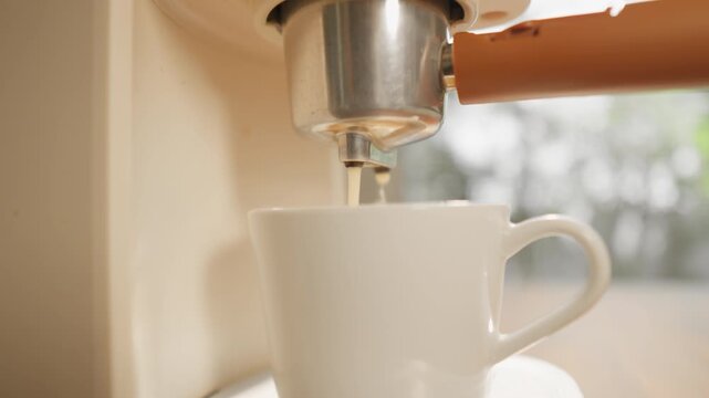 A close-up shot shows a coffee maker pouring hot coffee into a white coffee mug until it's full, with a soft, aromatic foam that's inviting to drink, in the warm morning light. The camera moves smooth