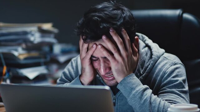 A stressed man sits at a desk with his hands on his face in front of a laptop.