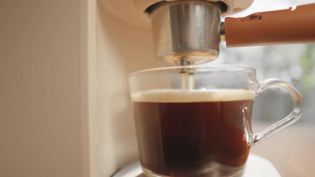 A close-up coffee maker brewing hot coffee into a clear glass mug, releasing a fragrant aroma and a smooth, inviting foam in the warm morning light.