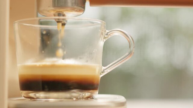 A close-up coffee maker brewing hot coffee into a clear glass mug, releasing a fragrant aroma and a smooth, inviting foam in the warm morning light.