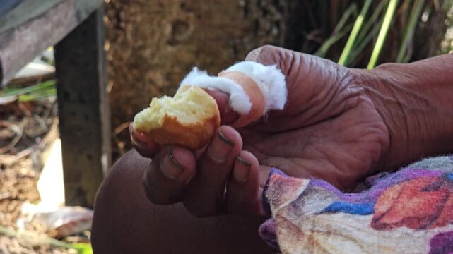 Close up of an elderly person with an injured bandaged finger experiencing tremor manula while holding a piece of bread and wearing a floral patterned shirt