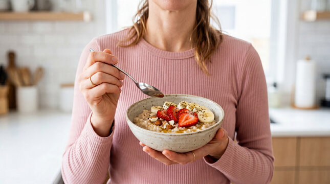 Woman holds bowl of oatmeal topped with fruit in a kitchen setting during morning hours