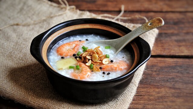 Rice gruel mixed with shimp and ginger, scallion slice and deep fried garlic and black pepper in black and brown bowl. ceramic spoon put back side on Sack and wood background