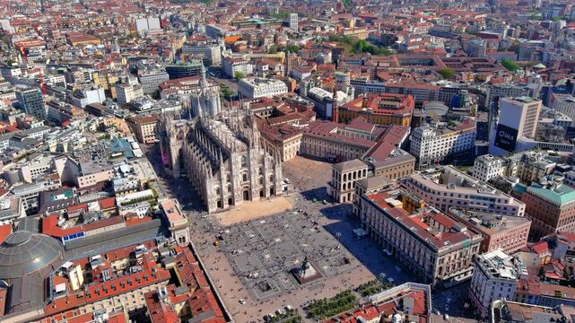 4K aerial drone view overlooking the magnificent Gothic architecture of the Duomo di Milano, the bustling Piazza del Duomo, and the historic surrounding cityscape in Milan, Italy.