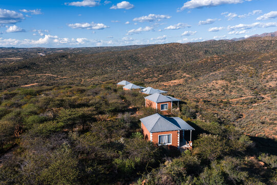Aerial small tin roof cabins line a ridgeline near Windhoek, Namibia, facing the Khomas Hochland in warm afternoon light with long shadows under puffy clouds.