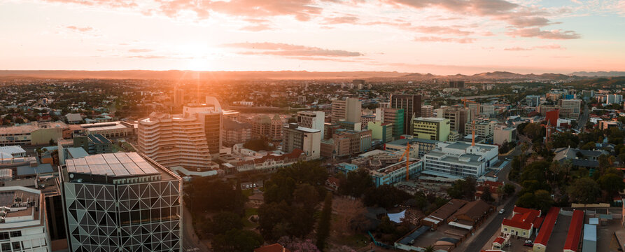 A wide aerial view of Windhoek, Namibia at sunset shows modern high rises, a steel framed building on the left, pastel towers, and arid hills and savanna beyond.