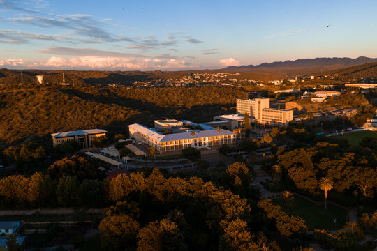 Aerial view of Windhoek, Namibia, at sunset shows modern institutional buildings, flat roofs, a glass and concrete complex, sports fields, and the Khomas Highlands.
