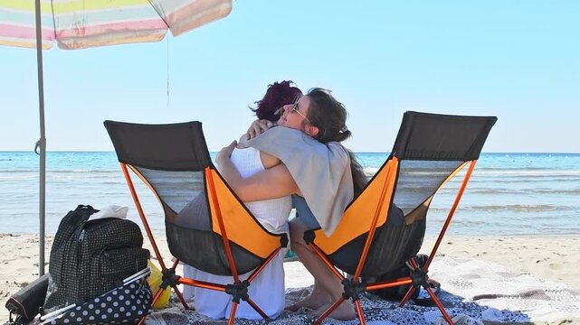 An older woman and a younger woman sharing a warm hug while relaxing in beach chairs by the seaside on a sunny day. intergenerational connection, tourism, summer accessories