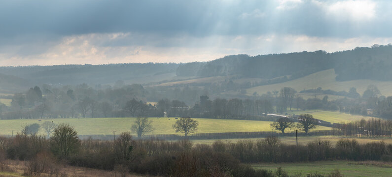 Misty Chilterns valley with crepuscular light rays over rolling farmland and wooded escarpment, Buckinghamshire, England, UK