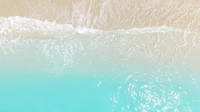 Static drone view from directly above showing crystal-clear turquoise water gently washing onto a sandy beach. Soft waves create natural patterns along the shoreline, highlighting the beauty