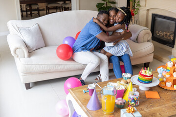 Obraz premium African American family hugging on sofa in living room around rainbow cake and balloons