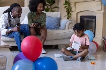 Obraz premium African American family celebrating at home child opening white gift box mom with phone, copy space
