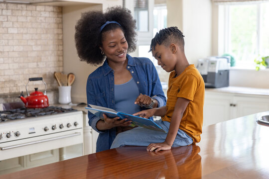 African American mother in denim shirt and son reading picture book on wooden countertop in kitchen