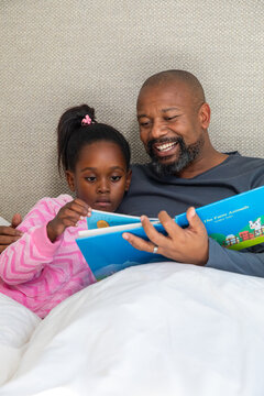 African American father and child reading Farm Animals book on bed, child wearing pink pajamas