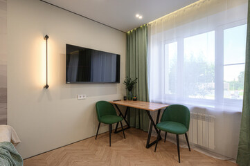 Fototapeta premium Modern corner setup with wooden table, two green upholstered chairs, wall-mounted TV, vertical LED light, layered curtains, and herringbone wooden floor