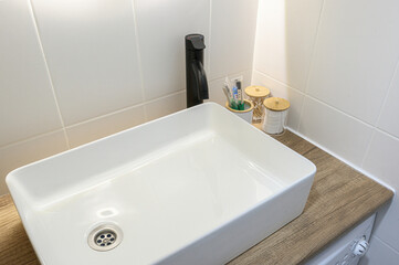 Fototapeta premium white rectangular vessel sink sits atop wooden countertop; paired with matte black faucet, against white tiled walls, with bamboo containers holding toiletries nearby