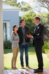 Real estate business market, house home building purchase sale. Two men and a woman standing outside of a house. The man on the right holds a clipboard, and the woman stands beside him.