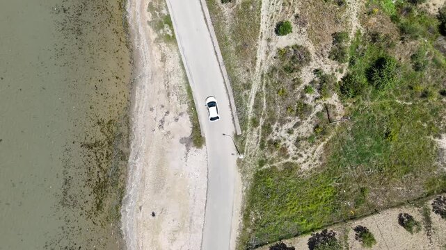 Aerial view of a white car driving along a winding rural road beside a tranquil pond, surrounded by lush greenery and patches of grass and trees