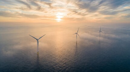Naklejka premium Offshore wind turbines standing in a calm open sea at sunrise with mist and dramatic clouds, renewable energy with copy space