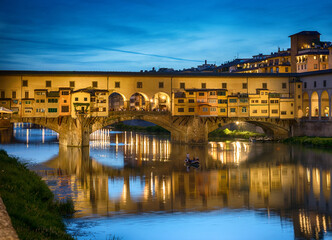 Night view of Florence, Italy, with the illuminated Ponte Vecchio bridge spanning the Arno River. Historic architecture, reflections on calm water, and a deep blue twilight sky  © Tryfonov