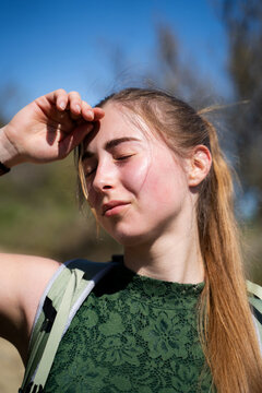 Exhausted woman backpacker shielding her face from intense sunlight while outdoors, standing in the sunlight with closed eyes and a tired expression on her faces, close-up portrait.
