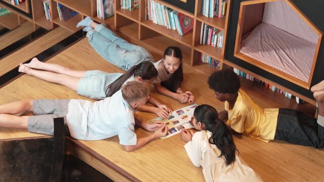 Group of diverse children lying down in circle while reading a book at library. Top view of girl sharing a magazine while pointing at interested topic and talking with lovely friends. Edification