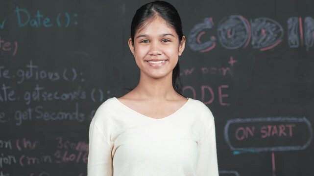 Caucasian woman looking at camera while standing at blackboard. Cute happy student smiling while stand in front of blackboard with engineering prompt or code in STEM technology class. Edification