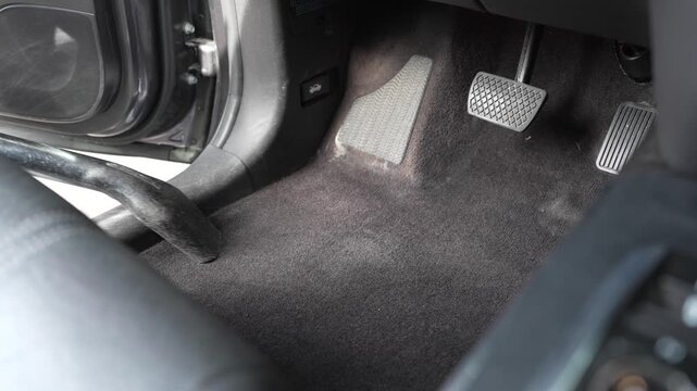 A medium close-up shot of a man's hand vacuuming the driver side carpet floorboard inside a vehicle. The video captures the cleaning process around the footwell area and pedals during automotive