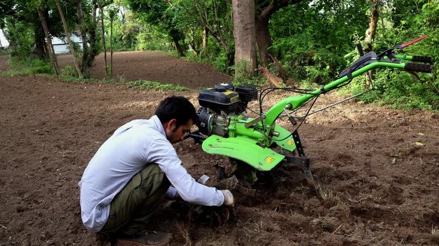 Closeup video of a young farmer cleaning grass stuck inside power tiller tractor cutting blades before starting agricultural field work during summer season in Himachal Pradesh, India.