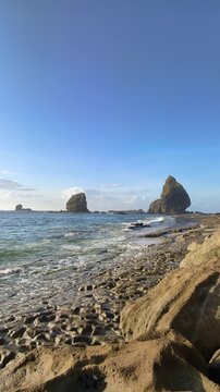 Stunning vertical view of rocky beach with clear blue ocean water and massive sea stack rock formations under a bright sky. Papuma Beach, Jember, Indonesia.