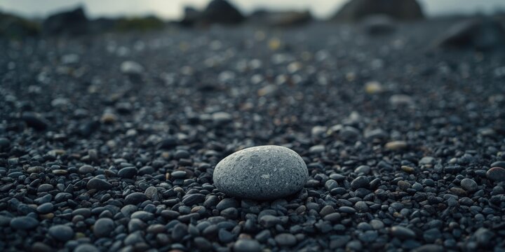 Single gray stone on coarse black pebble beach with indistinct water and stones in background and copy space