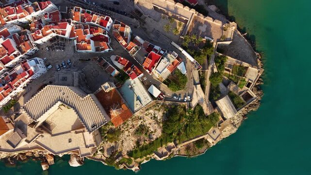 Top-down view of Peniscola castle complex featuring diamond-patterned church roof tiles, medieval fortifications and terracotta old town rooftops surrounded by turquoise Mediterranean Sea, Spain