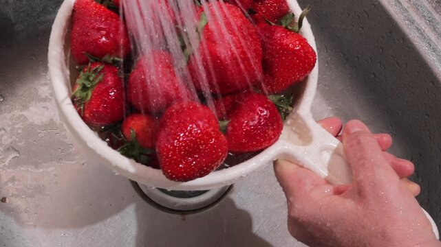 a woman washes ripe strawberries in a colander under the pressure of tap water over the sink
